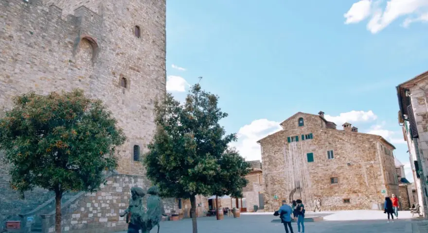 Piazza nel centro storico di Castellina in Chianti con torre medievale, edifici in pietra e scultura.