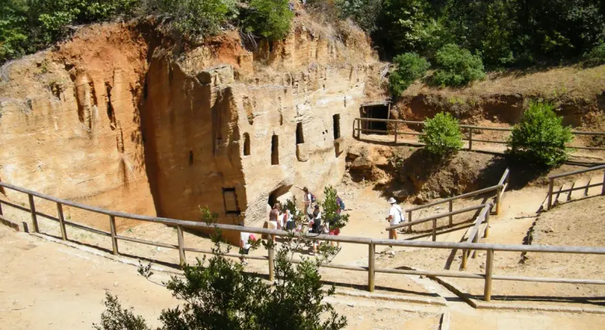 Antiche cave di tufo scavate nella roccia, con finestre e ingressi. Un gruppo di turisti esplora questo luogo affascinante, tra i **baratti cosa vedere** e la natura rigogliosa.