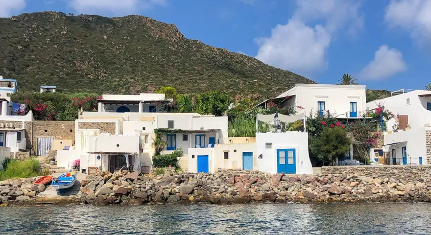 Case bianche con porte blu sull'isola di Panarea, con il mare calmo e una collina verde sullo sfondo.