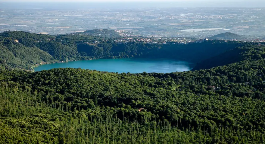 Vista panoramica del lago di Albano, circondato da rigogliosa vegetazione, con il borgo di Rocca di Papa in lontananza.