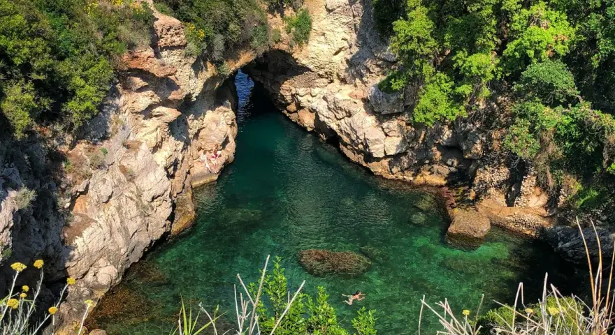Bagni Regina Giovanna: una baia nascosta con acque cristalline, rocce scolpite e vegetazione lussureggiante. Persone si godono il sole e nuotano.