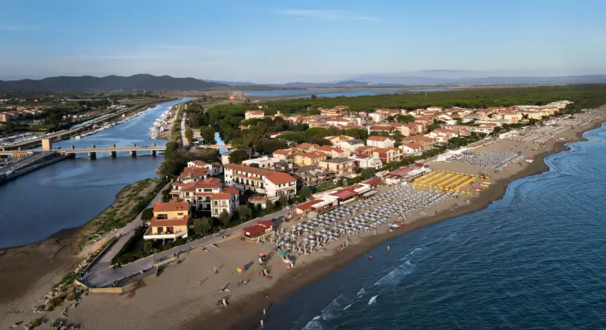 Vista aerea di Castiglione della Pescaia: un canale navigabile con barche, case con tetti rossi e una spiaggia attrezzata con ombrelloni e lettini, bagnata dal mare.