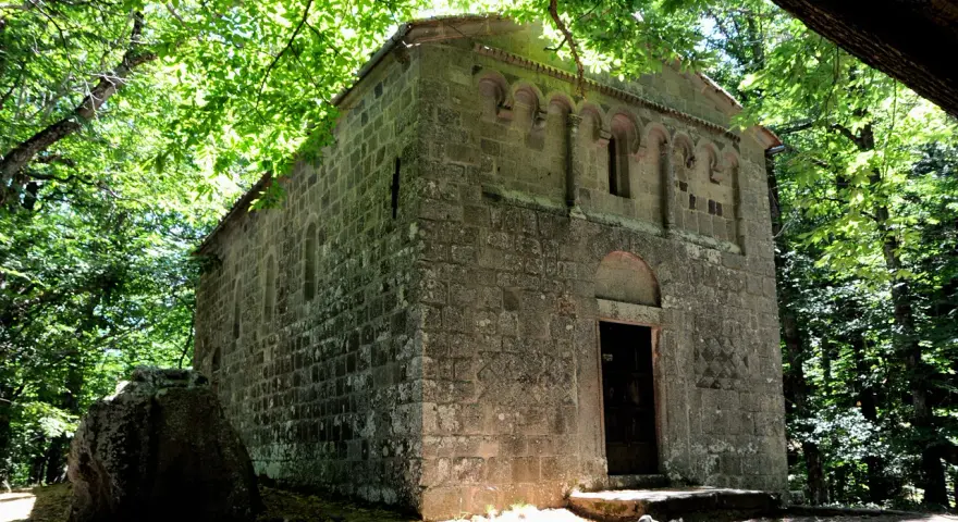 Antica pieve in pietra nel bosco Isabella, Radicofani. La luce filtra tra gli alberi illuminando la facciata.