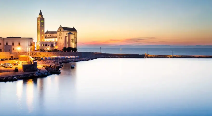 La cattedrale di Trani si erge maestosa sul mare al tramonto, illuminata da luci calde.