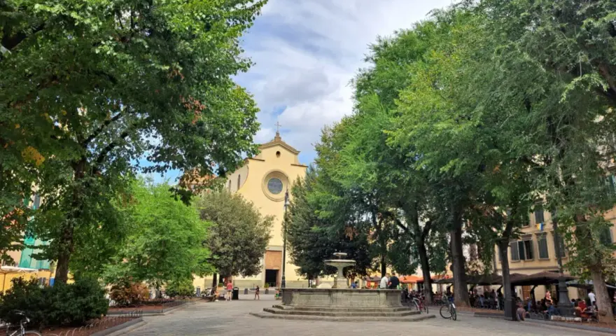 Piazza Santo Spirito a Firenze, con la chiesa sullo sfondo, una fontana al centro e alberi rigogliosi che incorniciano la scena.