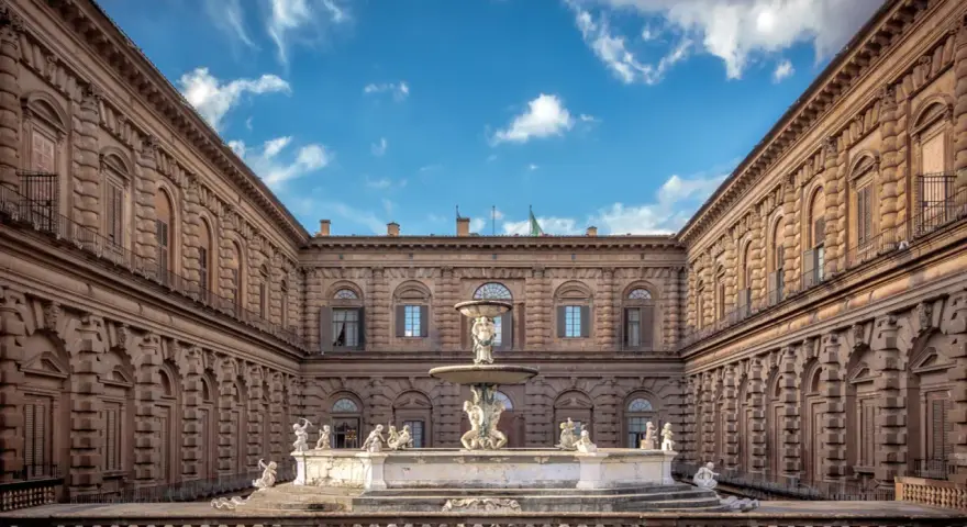 Cortile del Palazzo Pitti con fontana e statue, sotto un cielo azzurro con nuvole bianche.