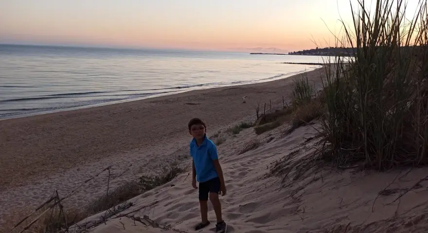 Bambino con occhiali su una duna di sabbia a Porto Palo, con il mare calmo e il cielo al tramonto sullo sfondo.