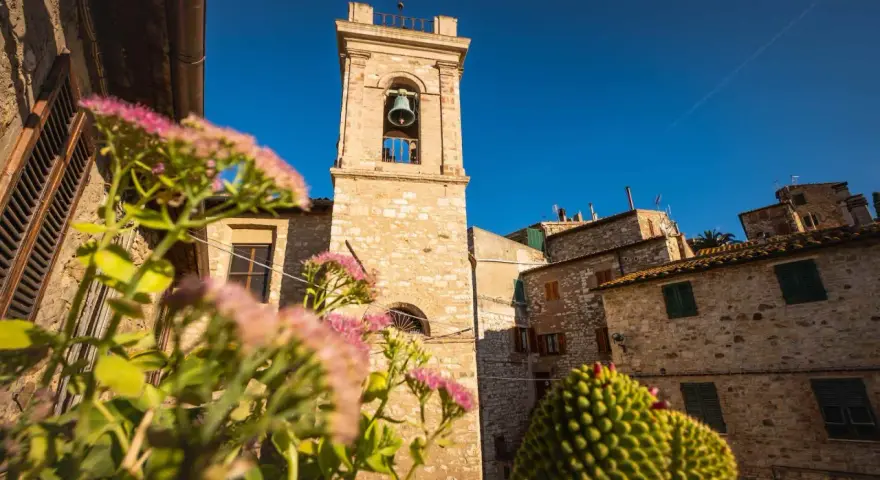 Campanile in pietra in un borgo delle Colline Metallifere, con fiori rosa in primo piano e cielo azzurro.