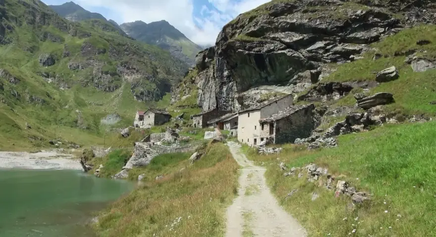 Antico borgo in pietra nel **Val di Susa**, con un sentiero che sale verso le case arroccate su un pendio verdeggiante, vicino a un lago alpino.
