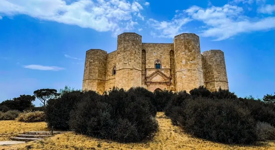 Castel del Monte, un'icona pugliese, si erge maestoso su una collina, circondato da una vegetazione rigogliosa sotto un cielo azzurro.
