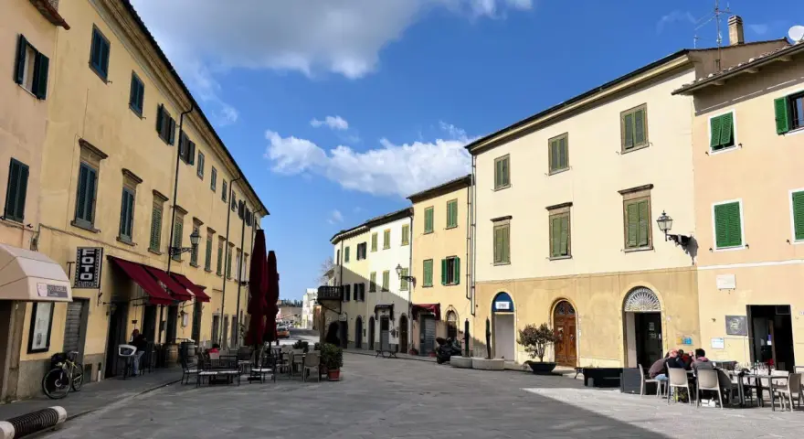 Piazza tranquilla a Pomarance, cosa vedere: edifici storici con persiane verdi, tavolini all'aperto di un bar.