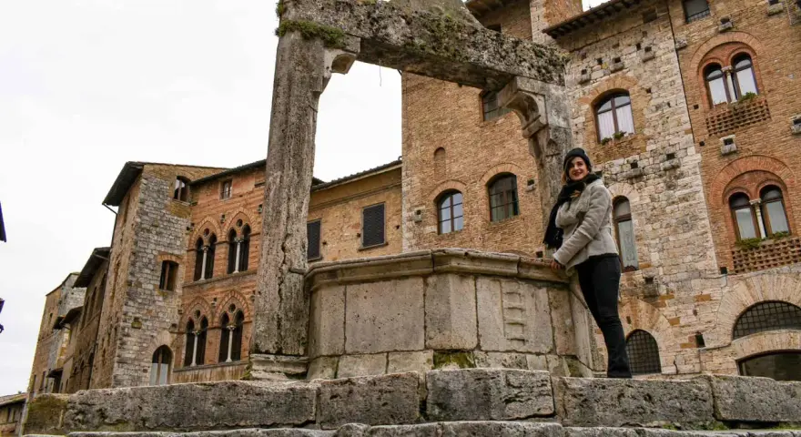 Donna sorride vicino all'arco in pietra e al pozzo di San Gimignano, con palazzi medievali sullo sfondo.