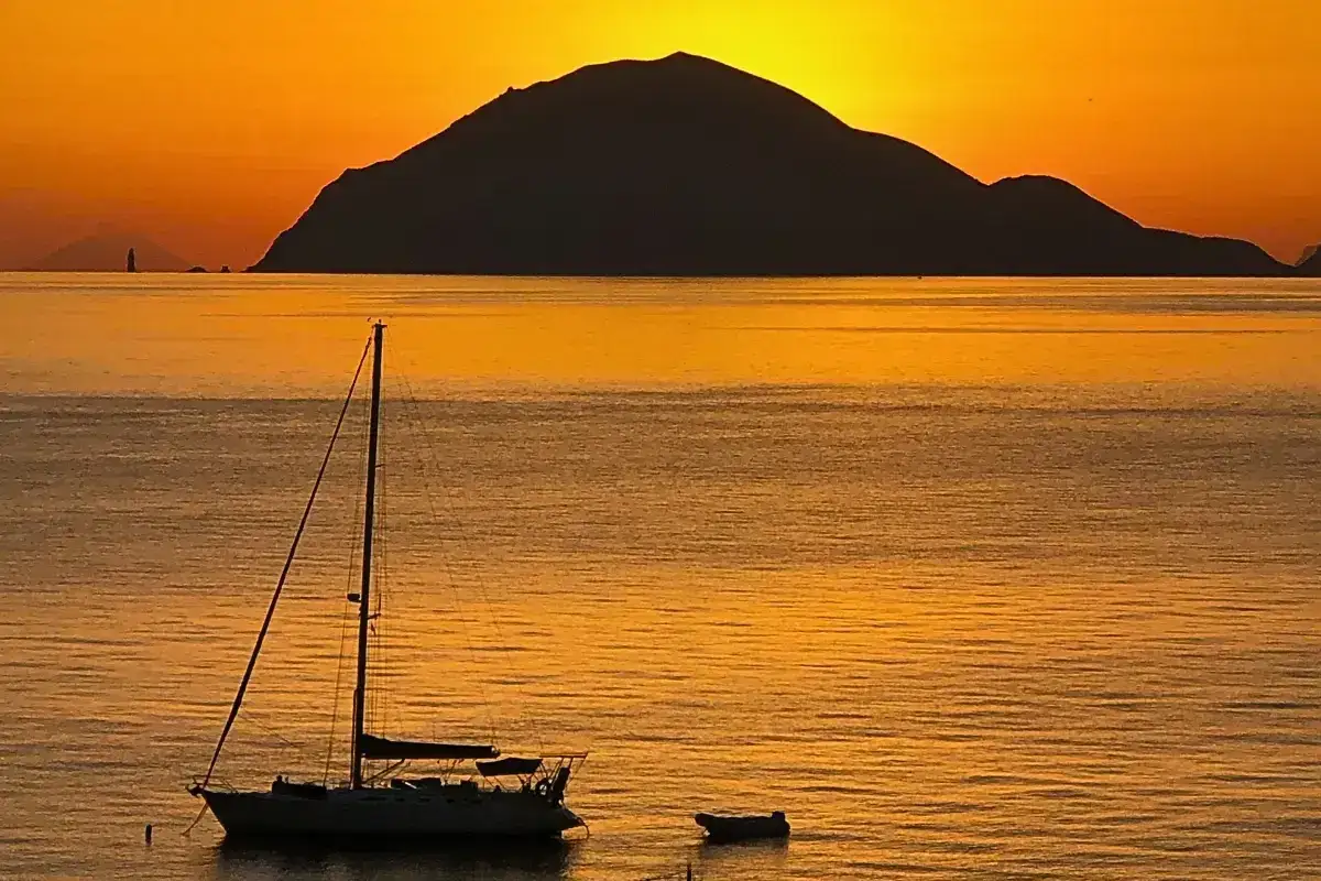 Tramonto dorato sulle Isole Eolie, con un veliero all'ancora e un faro in lontananza. Un'immagine che ispira a scoprire cosa vedere in questo arcipelago magico.