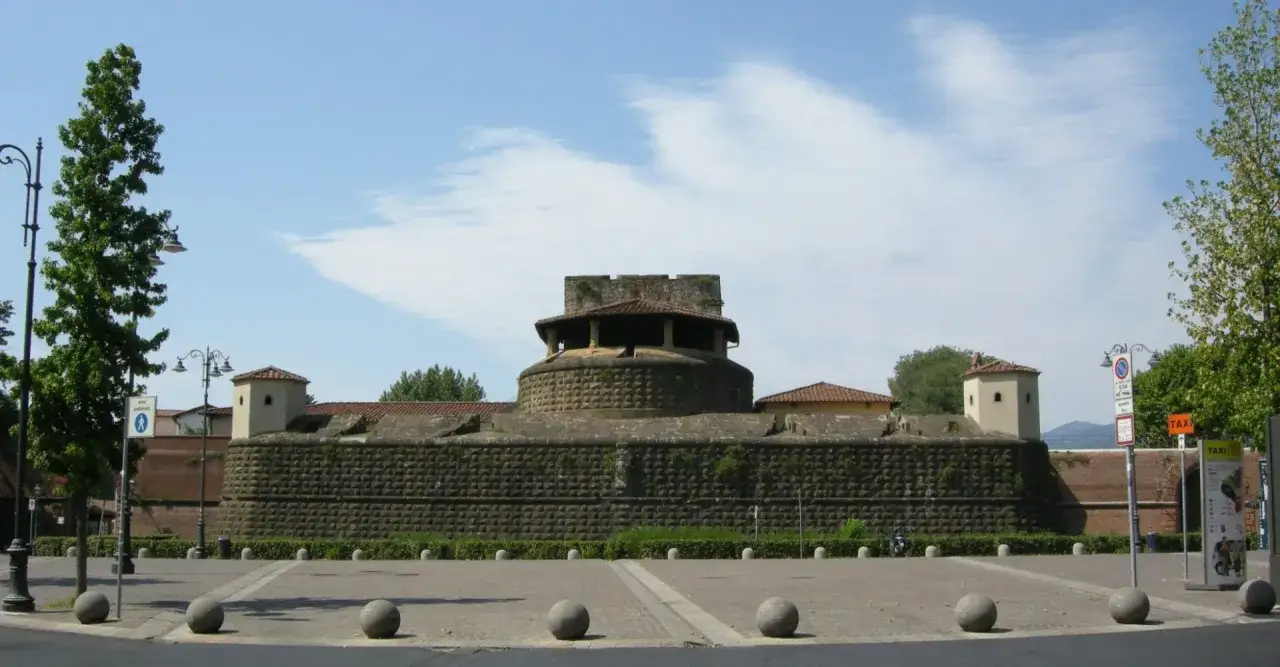 Imponente fortezza da basso con torri angolari e un torrione centrale circolare, circondata da un'area pedonale con lampioni e palle di pietra.