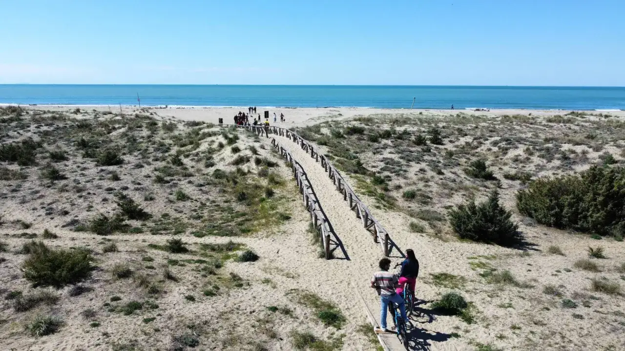 Coppia in bicicletta su passerella sabbiosa verso la spiaggia libera di Viareggio. Mare azzurro e cielo limpido.