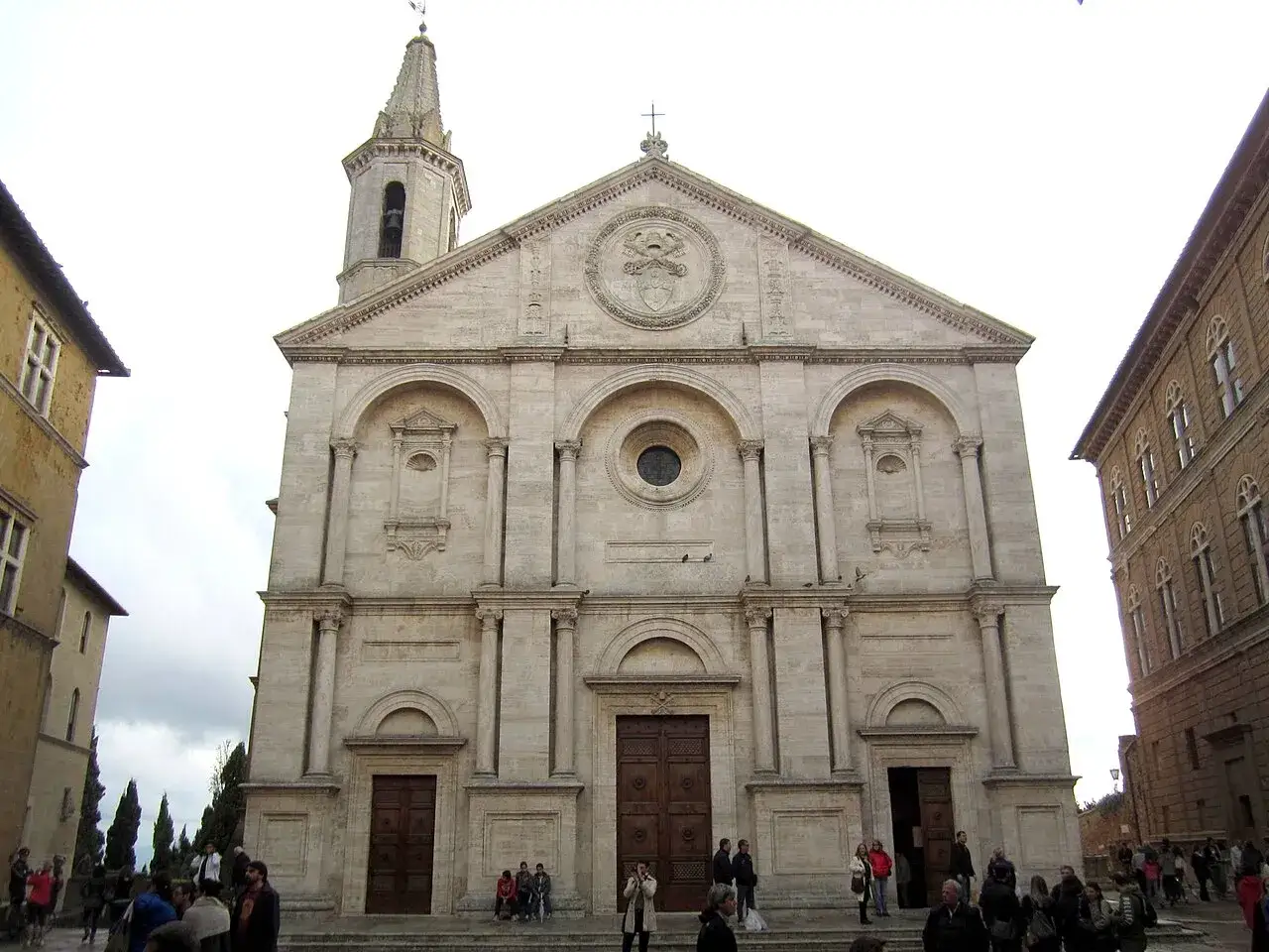 Il Duomo di Pienza, imponente facciata in travertino, con la sua torre campanaria e la piazza antistante animata da visitatori.