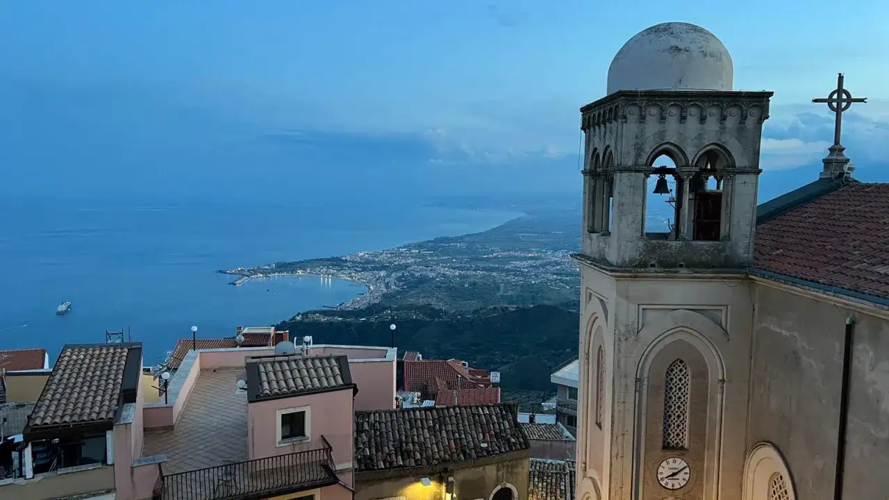 Vista panoramica di Castelmola cosa vedere: campanile di una chiesa, tetti e il mare al tramonto.