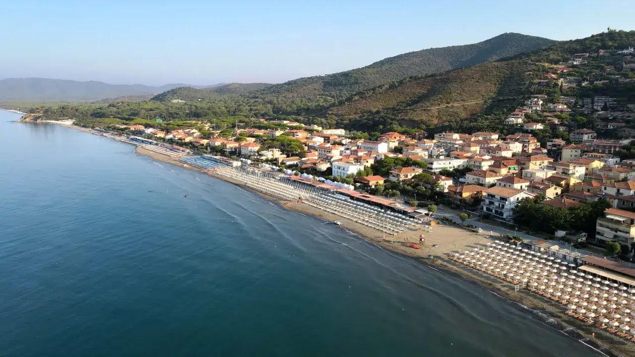 Vista aerea di Castiglione della Pescaia: un lungo tratto di mare azzurro lambisce una spiaggia attrezzata con file di ombrelloni bianchi, alle spalle un paese con tetti rossi e colline verdi.