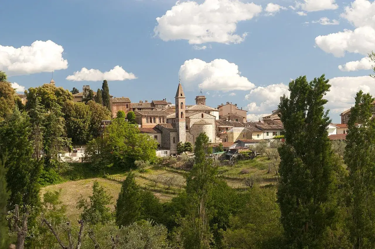 Panoramica di Castelnuovo Berardenga, un borgo toscano con un campanile svettante, circondato da verdi colline e alberi rigogliosi sotto un cielo azzurro con nuvole bianche.