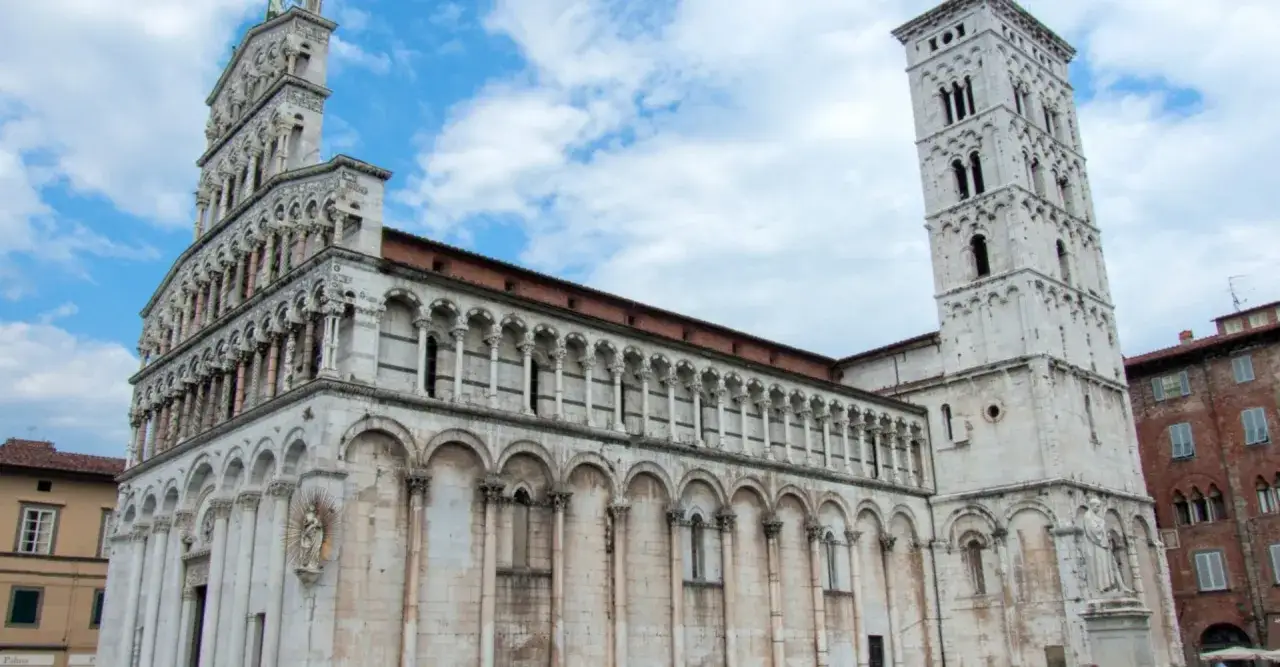 La splendida facciata della chiesa di San Michele in Foro a Lucca, con il suo campanile imponente, si staglia contro un cielo azzurro con nuvole bianche.