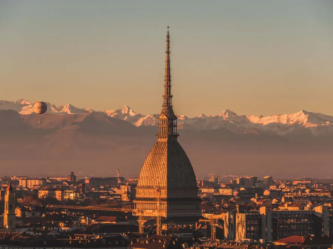 La Mole Antonelliana domina Torino al tramonto, con le Alpi innevate sullo sfondo e una mongolfiera che fluttua nel cielo dorato.