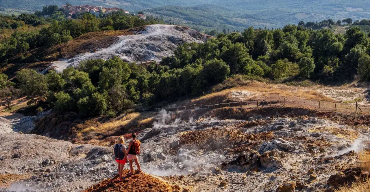 Due escursioniste ammirano la Valle del Diavolo, un paesaggio lunare con fumarole fumanti e rocce colorate, sullo sfondo di colline verdi e un borgo toscano.