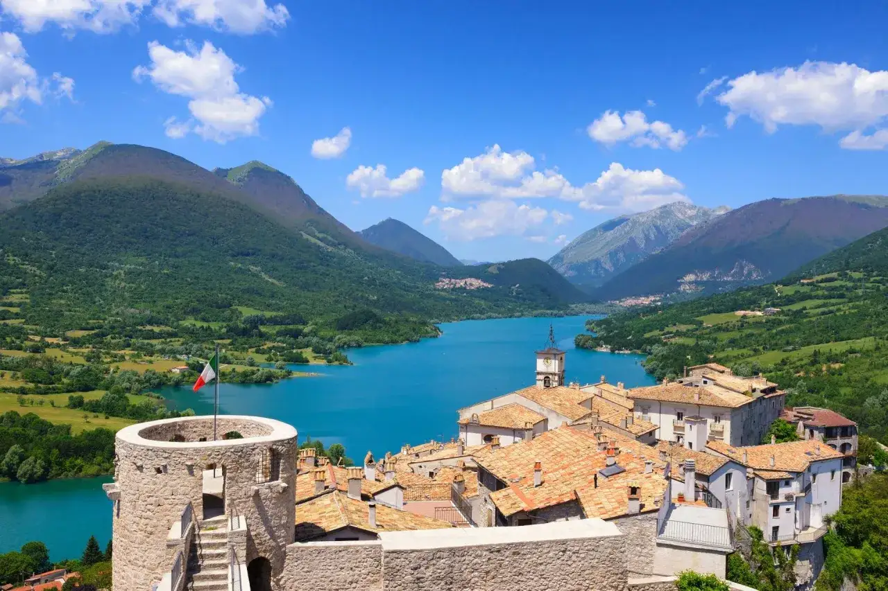 Villetta Barrea: cosa vedere? Un borgo con torre medievale e bandiera italiana domina un lago azzurro circondato da montagne verdi.