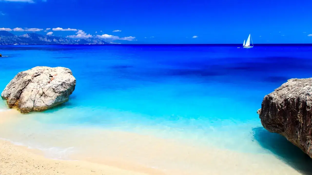 Spiaggia con rocce e mare cristallino, un veliero all'orizzonte. Un assaggio de le 10 spiagge più belle d'Italia.