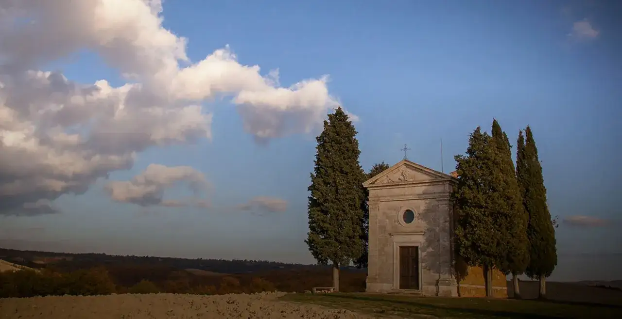 Piccola chiesa in pietra con cipressi, simbolo del Val d'Orcia, sotto un cielo azzurro con nuvole soffici.