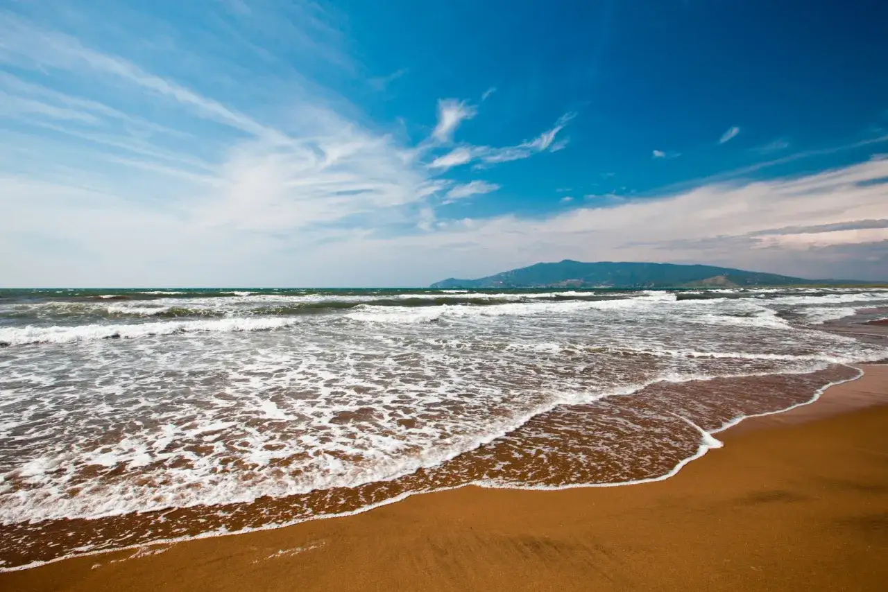 Onda che lambisce la spiaggia bianca di Ansedonia, con il mare mosso e le montagne all'orizzonte sotto un cielo azzurro e nuvoloso.