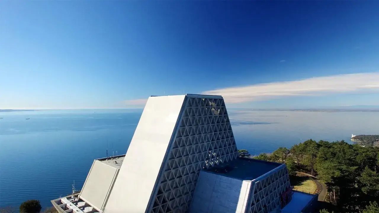 Il Santuario di Monte Grisa domina il mare azzurro sotto un cielo limpido. La sua architettura moderna, con la facciata a triangoli, si staglia contro l'orizzonte.