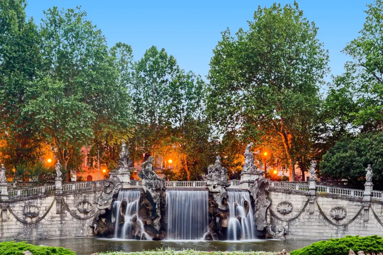 Fontana barocca con cascate nel Parco del Valentino, Torino. Statue e alberi verdi illuminati da luci calde.
