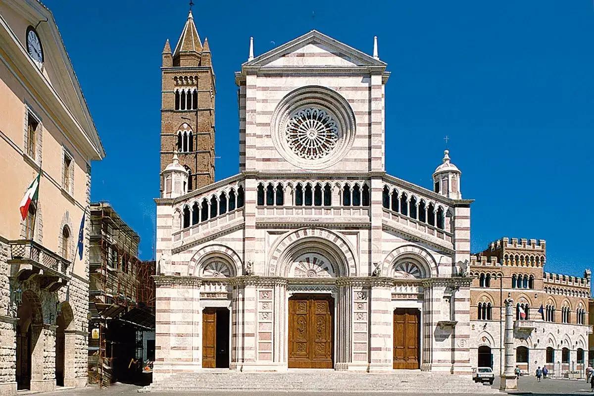 Il Duomo di Grosseto, con la sua facciata a strisce bianche e rosa, domina la piazza. Accanto, il campanile svetta contro il cielo azzurro.