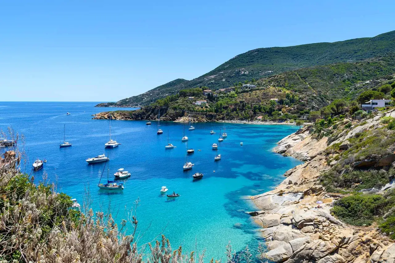 Baia con acque cristalline e barche a vela all'Isola del Giglio. Le spiagge sono incastonate tra scogliere rocciose e macchia mediterranea.