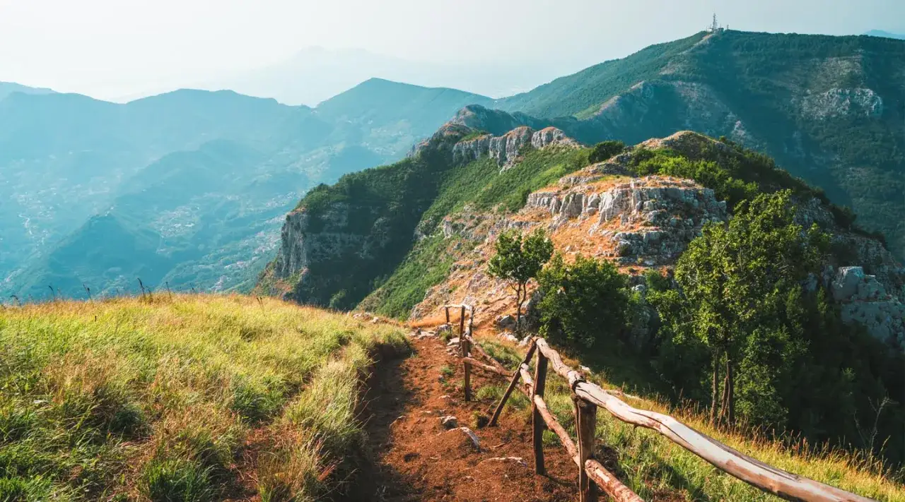 Sentiero sterrato con staccionata in legno sul valico di Chiunzi. Panoramica su montagne verdi e rocciose, con un villaggio in lontananza.