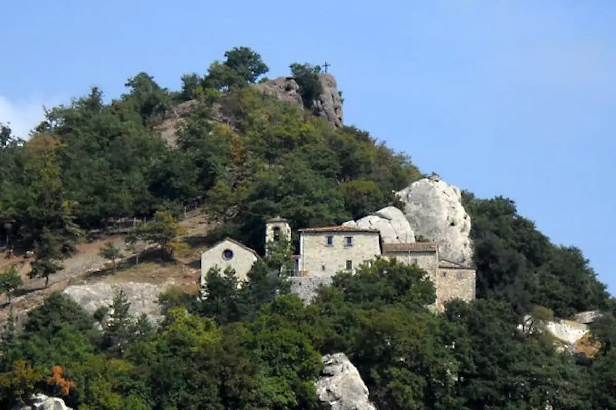 Antico eremo di Cerbaiolo, un complesso di edifici in pietra arroccato su una collina boscosa, con una croce in cima alla roccia.