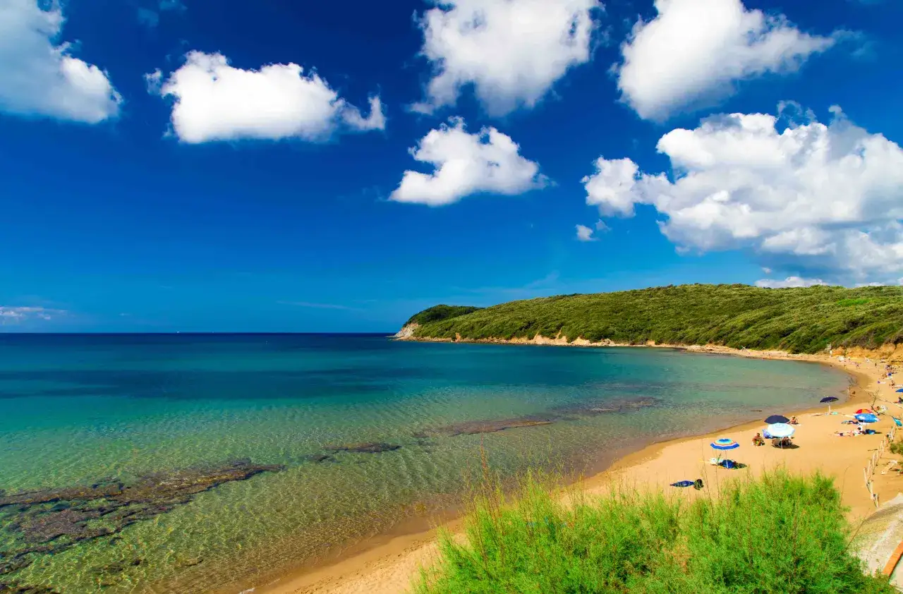 Spiaggia dorata e mare cristallino sulla costa degli Etruschi, con colline verdi e cielo azzurro punteggiato di nuvole bianche.