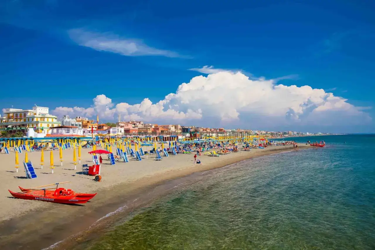 Spiaggia affollata a Ostia Mare, Roma, con ombrelloni gialli e blu, lettini, edifici colorati e il mare azzurro sotto un cielo limpido.