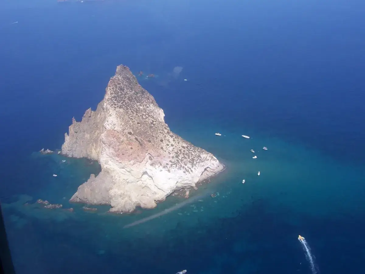 Vista aerea dell'isola di Panarea, con scogliere rocciose e acque cristalline. Diverse imbarcazioni solcano il mare blu intenso.