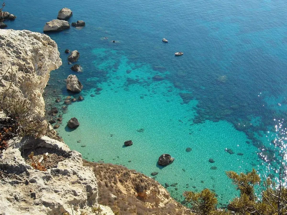 Scogliera rocciosa affacciata su acque cristalline e turchesi, con piccole imbarcazioni. Un angolo di paradiso che ricorda la spiaggia del Poetto.