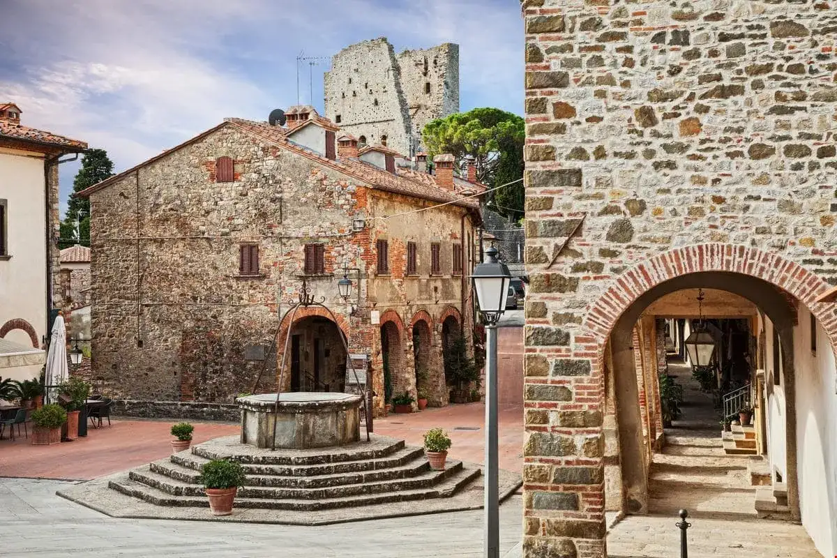 Antica piazza con pozzo a Civitella in Val di Chiana. Edifici in pietra e mattoni, un arco in primo piano e rovine di un castello sullo sfondo.