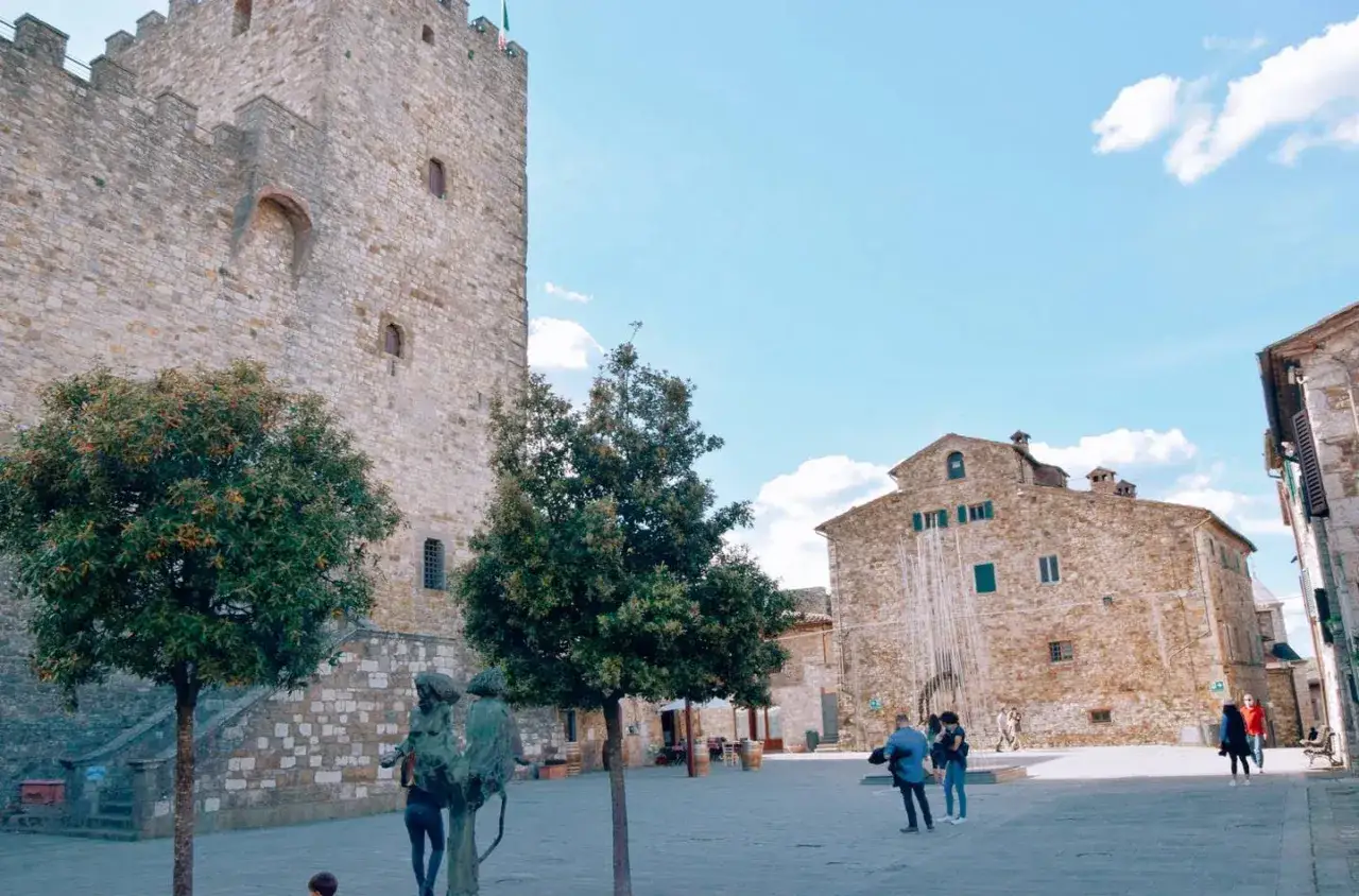 Piazza nel centro storico di Castellina in Chianti con torre medievale, edifici in pietra e scultura.