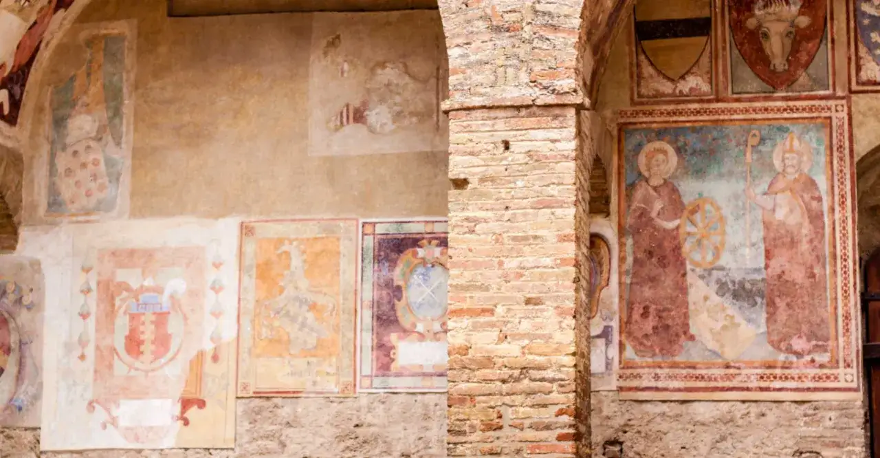 Affreschi medievali nel duomo di San Gimignano: stemmi araldici e figure sacre su muri antichi.