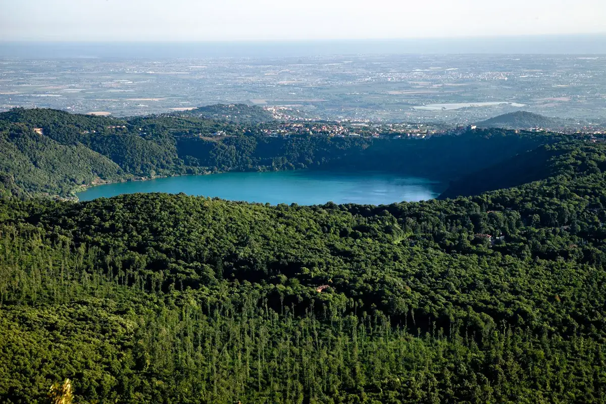 Vista panoramica del lago di Albano, circondato da rigogliosa vegetazione, con il borgo di Rocca di Papa in lontananza.
