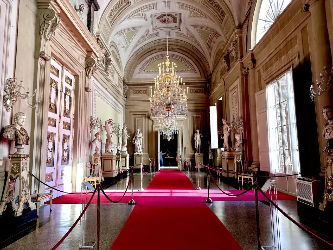 Corridoio sfarzoso di Palazzo Pitti con tappeto rosso, statue e un lampadario imponente.