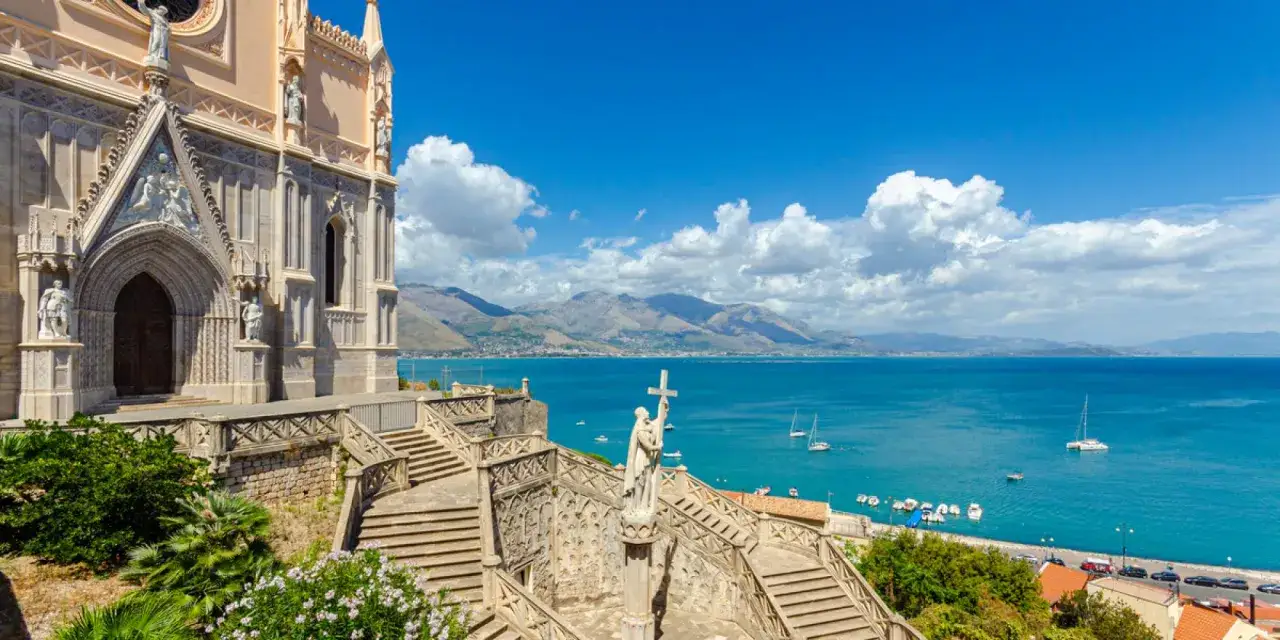La chiesa domina il mare azzurro della Riviera di Ulisse. Scale imponenti conducono al portale, mentre barche e montagne incorniciano il panorama.