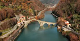 Il **ponte della Maddalena**, con i suoi archi irregolari, attraversa un fiume circondato da colline autunnali e case colorate.