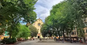 Piazza Santo Spirito a Firenze, con la chiesa sullo sfondo, una fontana al centro e alberi rigogliosi che incorniciano la scena.