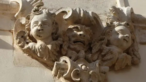 Dettaglio scultoreo barocco a Martina Franca: volti di putti e una maschera grottesca emergono dalla pietra calcarea, testimoni di un'antica arte.