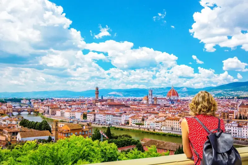 Vista mozzafiato di Firenze dal Piazzale Michelangelo. Una donna ammira il Duomo e il Ponte Vecchio sotto un cielo azzurro con nuvole bianche.